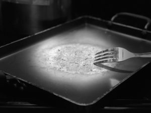 A black and white photo of a shiny stainless steel fork scraping the teflon coating off of a large rimmed rectangular griddle.
