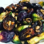 deep purple, caramelized black grapes, halved and mixed with bright green cucumber slices, and garnished with toasted sunflower seeds and tiny ribbons of fresh green basil. Served in a white bowl.