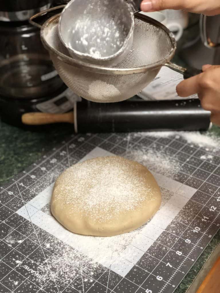 A disk of raw dough on top of grey and white measuring mat, with flour being sprinkled on top through a sieve. A black rolling pin is resting in the background.