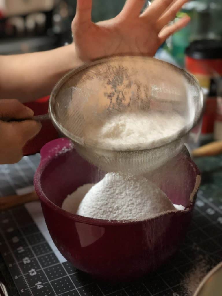 Flour being sifted through a sieve into a magenta-coloured plastic mixing bowl