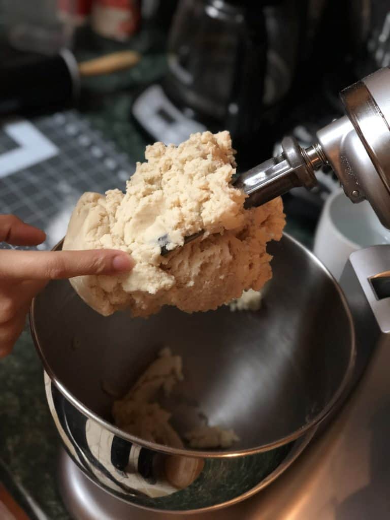A finger pressing into the finished cookie dough as it hangs on the lifted paddle attachment of an electric stand mixer with the silver mixing bowl below it. 
