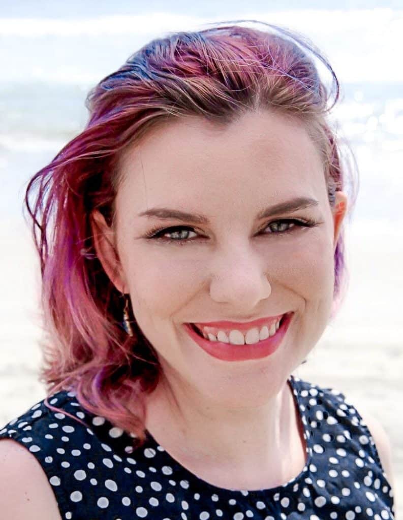 Close up photo of a white woman with magenta hair and a polka dot top, smiling at the beach