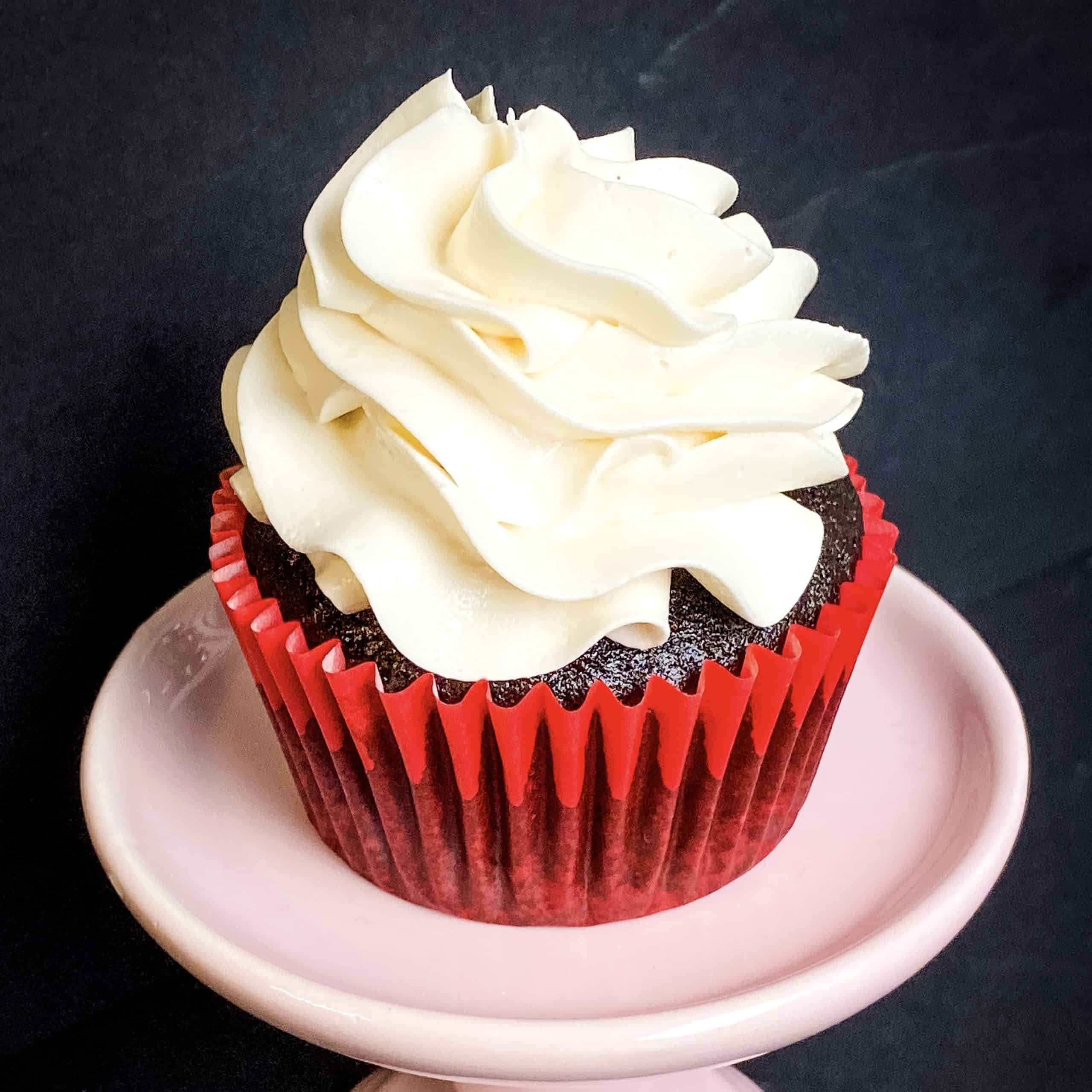 a cupcake with vanilla buttercream swirlled on top. it has a red case and is sitting on a pink cupcake stand with a black background.