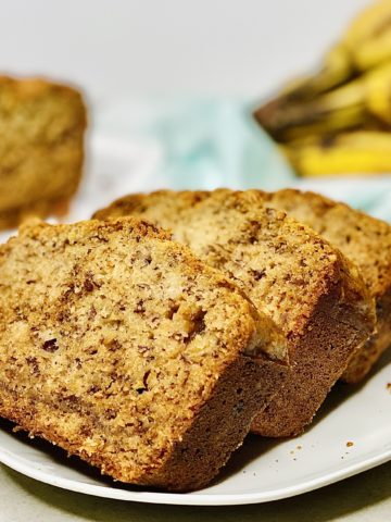 three slices of banana bread on a white plate, with ripe bananas and a sliced loaf in the background