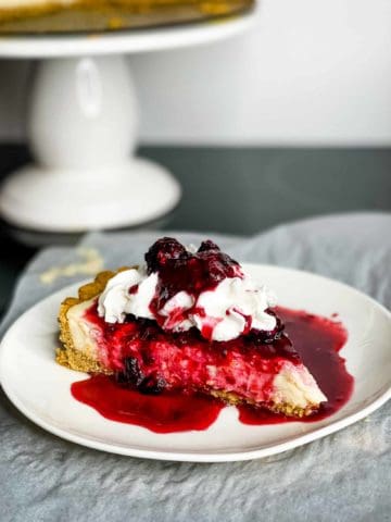 a slice of cheesecake topped with red berry sauce and whipped cream, on a white plate