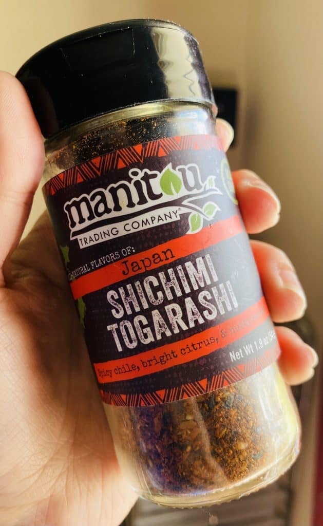 A spice bottle full of togarashi seasoning in a white woman's hand. It is the brand manitou trading company and reads natural flavors of japan shichimi togarashi spicy chile, bright citrus, and nutty sesame