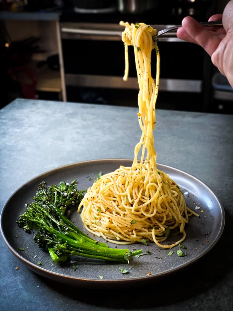 A view of a grey plate on a slate countertop. The plate has broccolini on one side and a mount of spaghetti on the other. A fork is lifting some of the spaghetti high into the air.