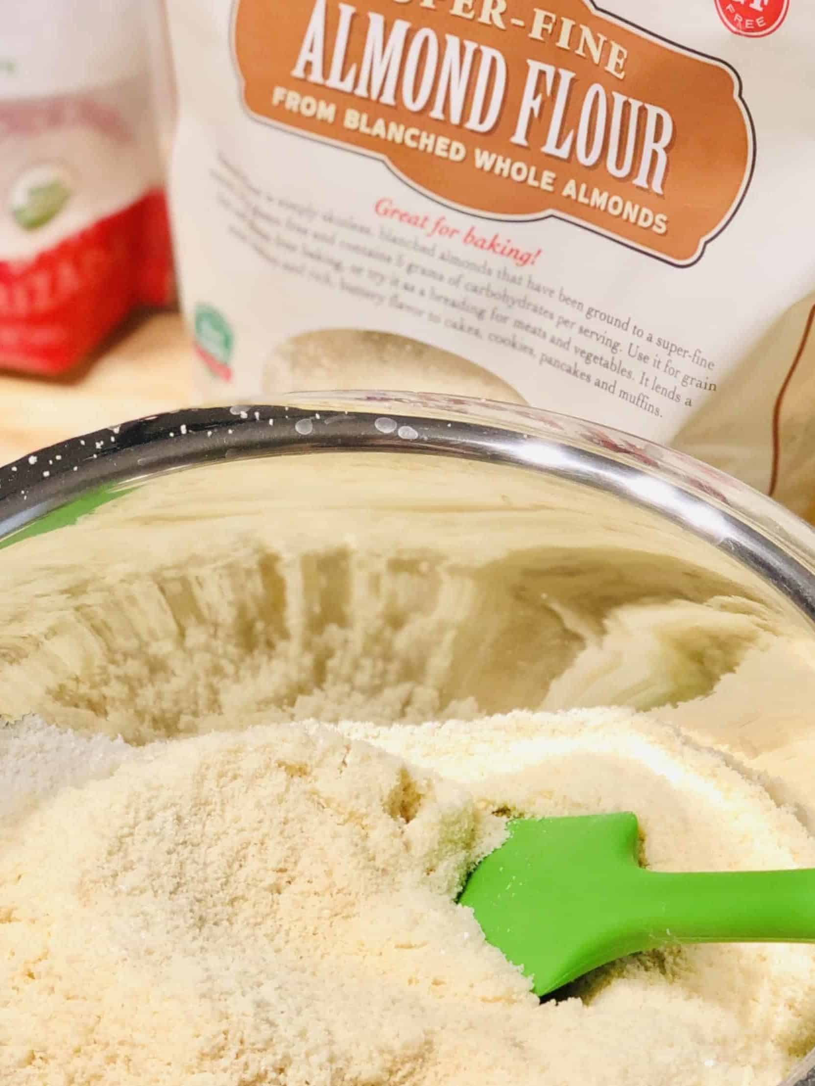 A stainless steel mixing bowl full of almond flour with a green silicone spatula in it. A bag of almond flour sits behind it.