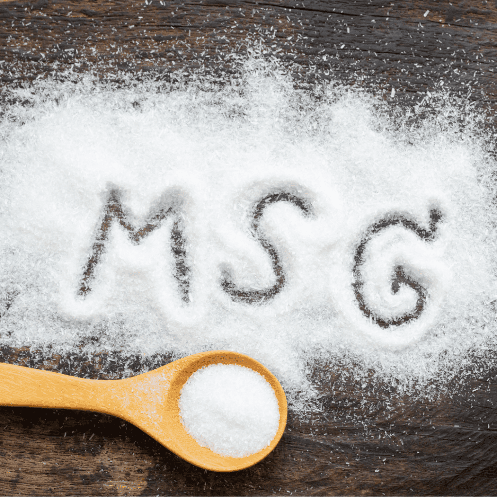 white powder sprinkled on a wooden tabletop with 'msg' written in it with a finger. A wooden spoon is on the bottom of the photo with More of the white powder in it.