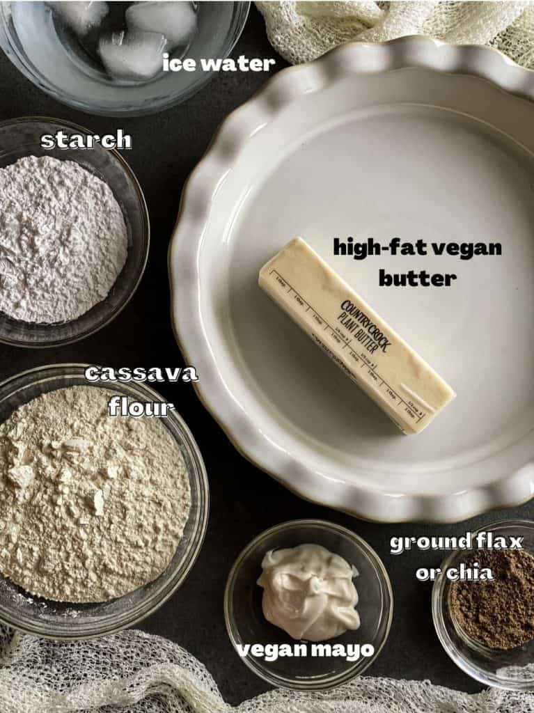 Ingredient shot for the gluten free pie crust. A stick of butter is in the white ceramic pie plate, surrounded by bowls filled with: ice water, starch, cassava flour, mayonnaise, and ground lax seed