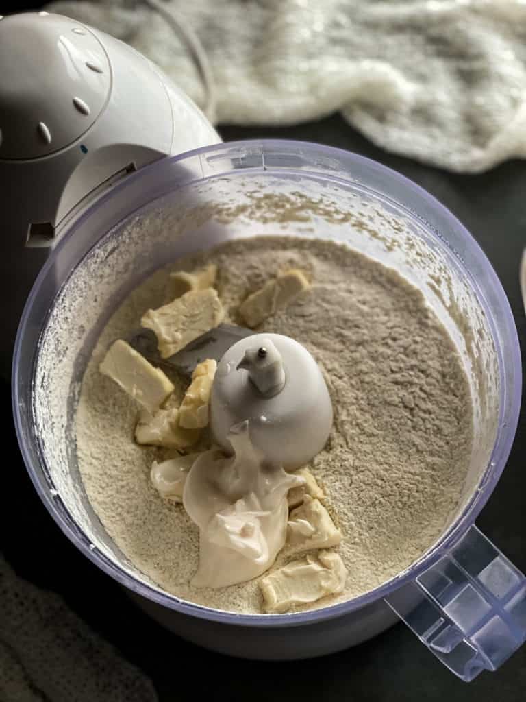 cubes of butter and dollop of mayonnaise sitting on top of the flour mixture in the bowl of the food processor