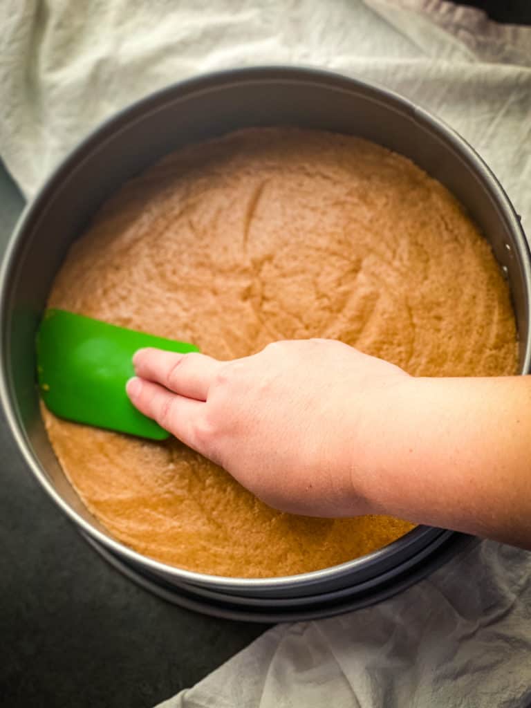 Pressing the dough down into the springform pan with a green silicone spatula. A white woman's hand is pressing it down with two fingers into the corners.
