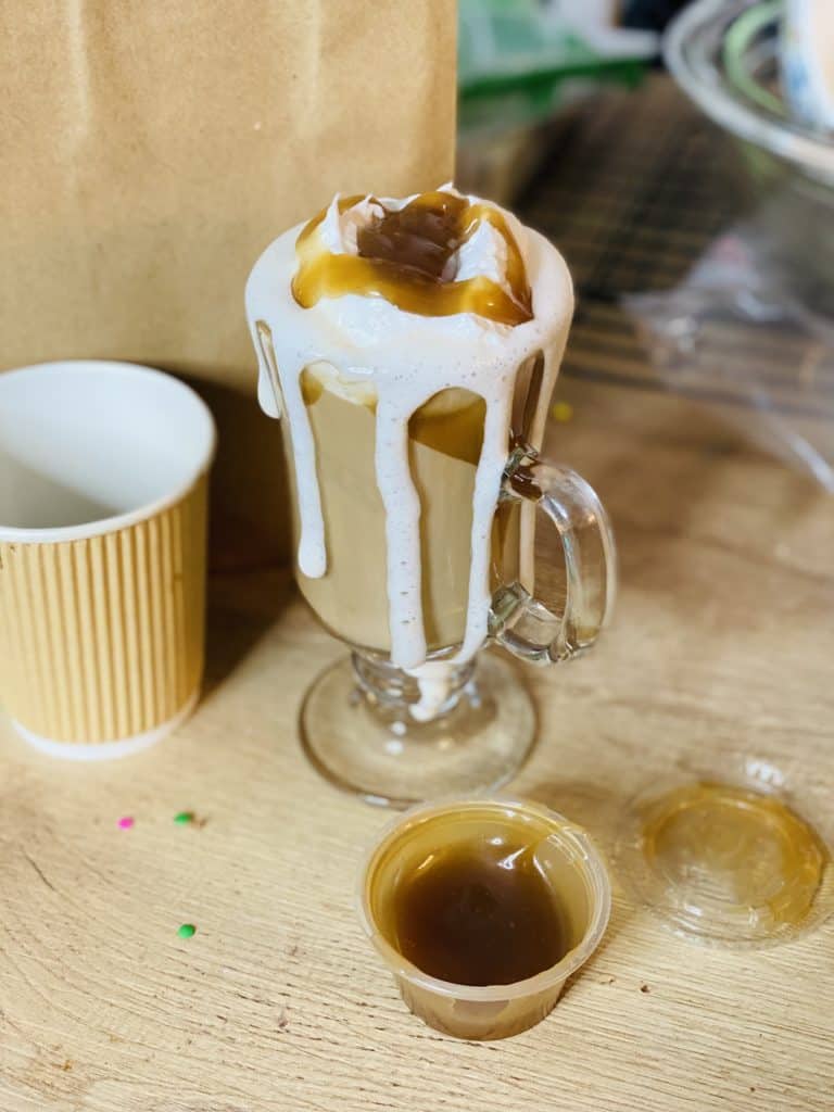 A tall coffee in a glass mug with lots of whipped cream and caramel sauce, with a container of caramel and empty paper coffee cup sitting next to it.