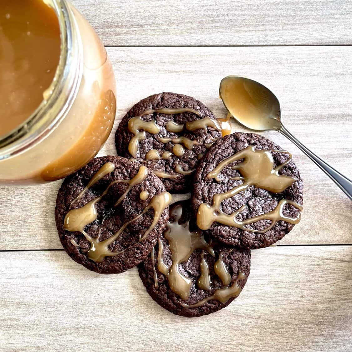 Four chocolate cookies drizzled with caramel sauce, next to a jar of caramel and a caramel covered spoon.