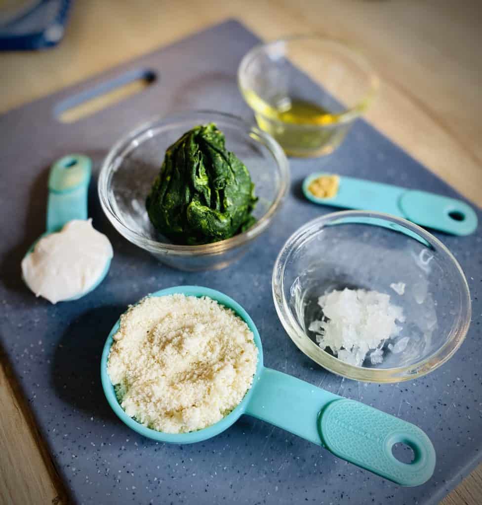 Olive oil, minced onion, a ball of squeeze-dried spinach, parmesan, minced garlic, and mayonnaise in various containers atop a blue cutting board.