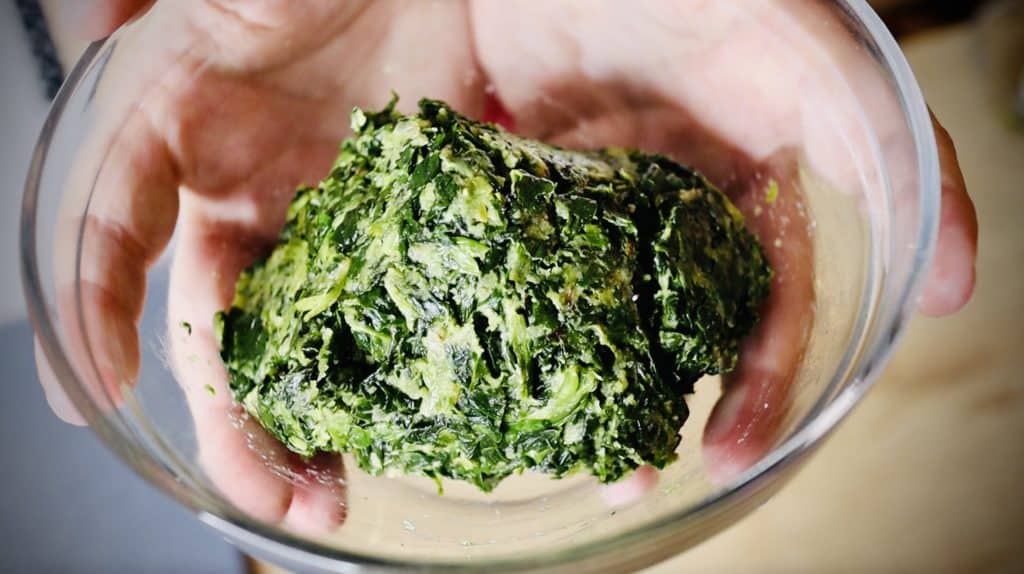 Two hands holding a clear glass bowl with a ball of spinach filling in it