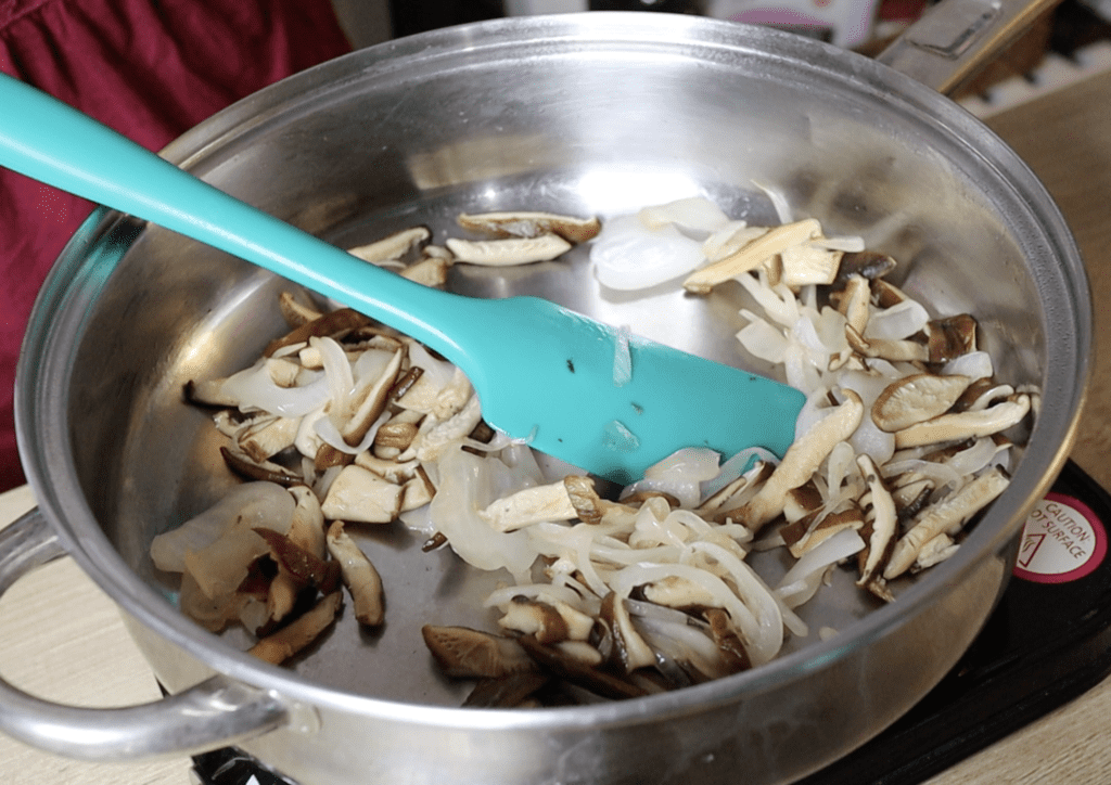 Sautéing onion and shiitake slices in a stainless steel skillet with a blue spatula 