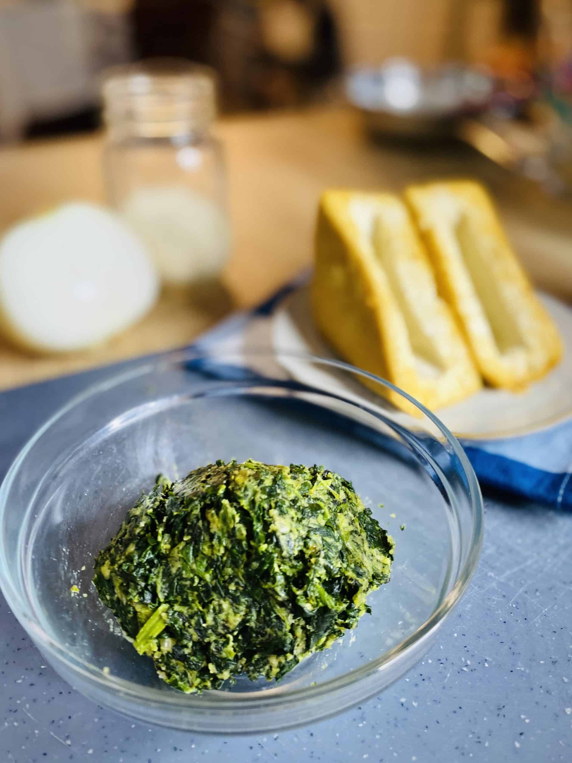 A clear glass bowl with a ball of spinach filling in it, sitting on top of a blue board. Blurred behind it are two empty tofu pockets, a cut onion, and jar of parmesan.