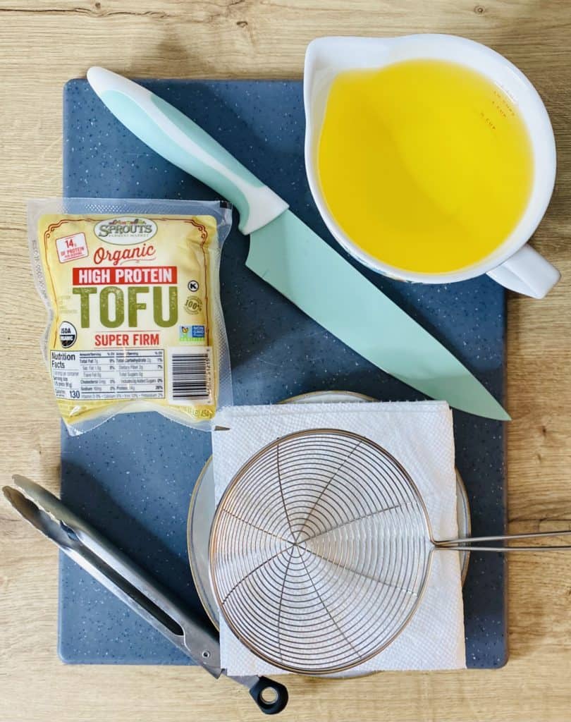A package of unopened tofu, a measuring cup with oil, a teal chopping knife, a pair of metal tongs, and a metal spider tool on top of a paper towel lined white plate, all of which are sitting on top of a blue cutting board on a wooden countertop.