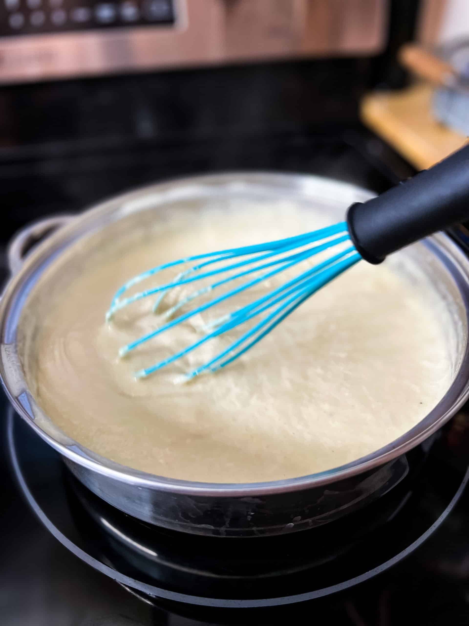a turquoise colored whisk stirring Alfredo sauce in a stainless steel pan
