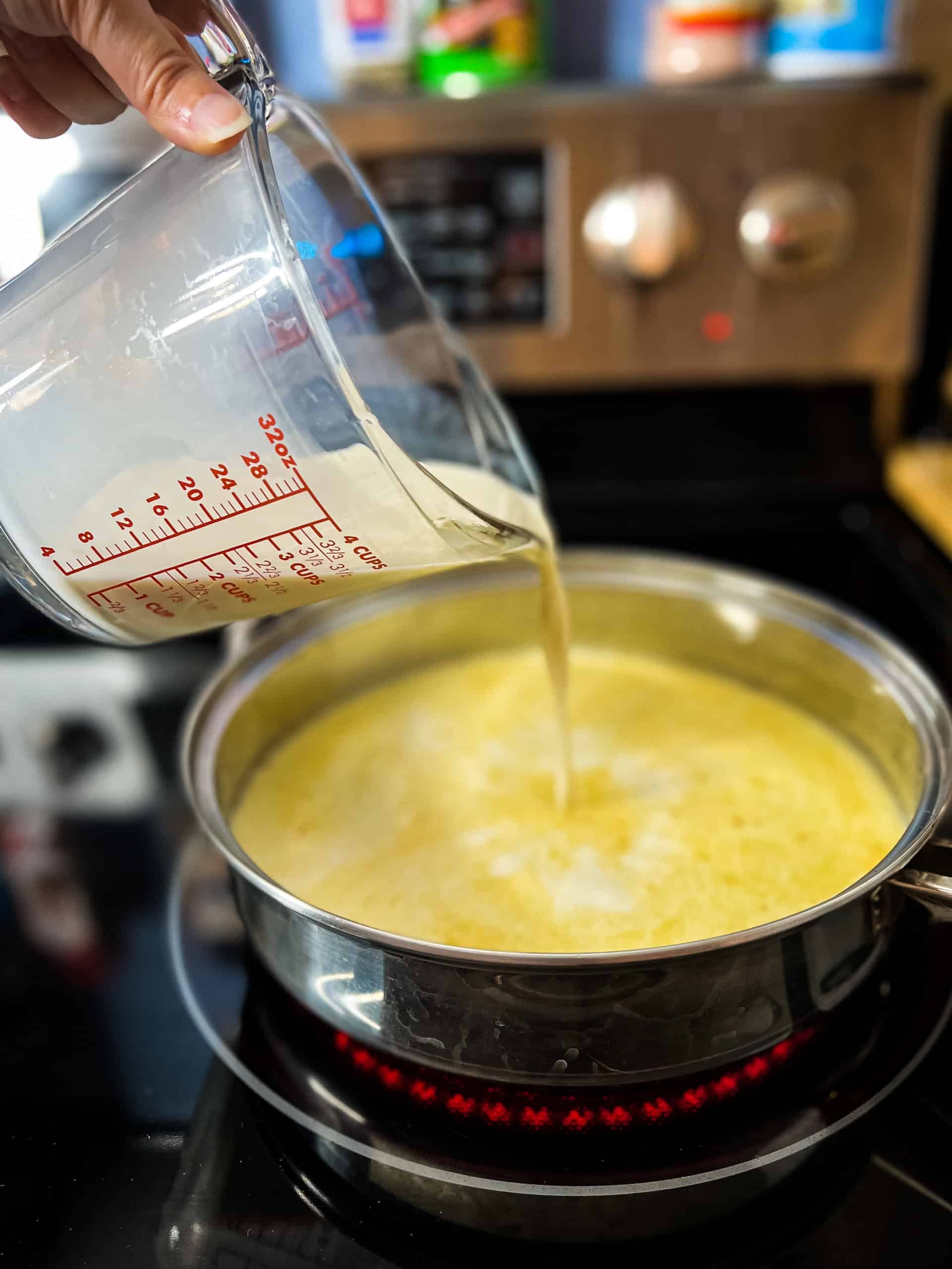 pouring milk from a 4c glass measuring cup into a stainless steel pan with melted butter
