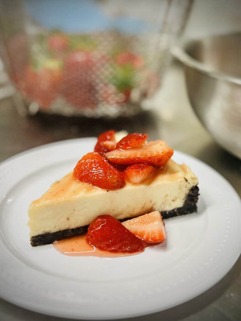 A slice of cheesecake with a chocolate cookie crust and macerated strawberries on top 